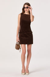 A woman is wearing a sleeveless, dark brown dress with a fitted top and a matching skirt that ends above the knees. She has on large, dark sunglasses and beige high-heeled shoes with a subtle pattern. She is holding a small beige handbag in her left hand.